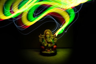 A colorful, glowing light painting swirls around a small statue of a deity with an elephant head, which is seated and adorned with intricate details. The lights create a dynamic and energetic pattern above the statue, illuminating it from above and highlighting its features.