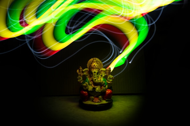 A colorful, glowing light painting swirls around a small statue of a deity with an elephant head, which is seated and adorned with intricate details. The lights create a dynamic and energetic pattern above the statue, illuminating it from above and highlighting its features.