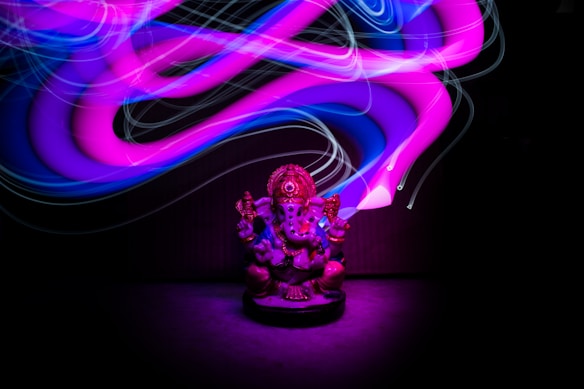 A statue of Ganesha sits in the foreground, surrounded by vibrant, swirling light trails in shades of purple, pink, and blue, creating a dynamic and mystical atmosphere.