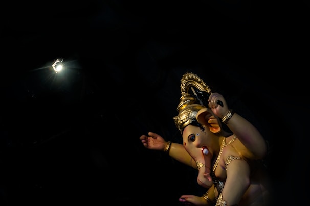 A statue of the Hindu deity Ganesha, adorned with gold jewelry and a headdress, stands against a dark background. The statue is illuminated by a spotlight, highlighting its intricate details and vibrant colors.