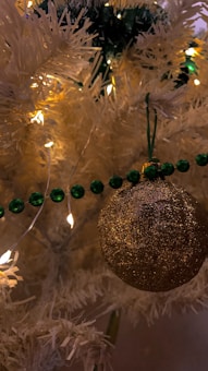 A close-up view of a decorated white artificial Christmas tree featuring a glittery gold ornament hanging from a green string. The tree is adorned with green beads and small warm white fairy lights adding a festive glow.