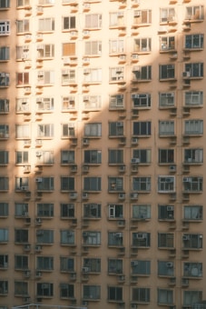 A high-rise apartment building with multiple uniform windows, each equipped with individual air conditioning units. The facade of the building is bathed in soft golden sunlight, casting geometric shadows across the surface.