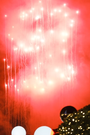 Bright fireworks illuminate the sky, with trails of sparks streaming downward against a red and smoky background. In the lower part of the image, there are festive decorations with lights and trees, creating a vibrant and celebratory atmosphere.
