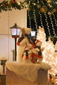 An enchanting Christmas scene features three white polar bear figures wearing red and black scarves, paired with a small Santa Claus figure. They stand on a snow-covered platform surrounded by festive decor, including a decorated white Christmas tree illuminated by warm lights. Above, a garland with gold baubles and fairy lights hangs, contributing to the festive atmosphere.