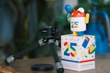 A figurine resembling an Android robot stands on a colorful box featuring the number 25 and various tech-themed icons. The robot is holding a blue balloon and wearing a white cap with a circular logo. Next to it, there is a black FUJIFILM camera tripod on a wooden surface, with a blurred background of greenery.