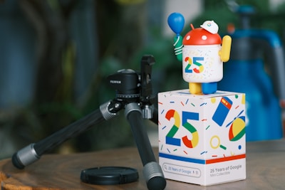 A figurine resembling an Android robot stands on a colorful box featuring the number 25 and various tech-themed icons. The robot is holding a blue balloon and wearing a white cap with a circular logo. Next to it, there is a black FUJIFILM camera tripod on a wooden surface, with a blurred background of greenery.