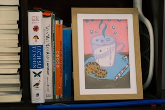 A framed illustration of a steaming cup with polka dots placed on a surface with cookies and a straw beside it. The background is pink with blue polka dots. This sits on a bookshelf among several books, including titles in different languages, with a small section of a sports racquet visible on the side.