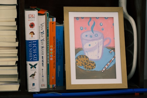 A framed illustration of a steaming cup with polka dots placed on a surface with cookies and a straw beside it. The background is pink with blue polka dots. This sits on a bookshelf among several books, including titles in different languages, with a small section of a sports racquet visible on the side.