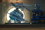 Two intricately designed robot figures engage in a dynamic pose within a glass display case. The left figure is predominantly white with turquoise accents, holding a large spear and shield, and features an orange element on its head. The right figure is primarily blue and gold, wielding a sword, poised in a combat stance. The background is illuminated by a bright circular light, creating a dramatic silhouette effect.