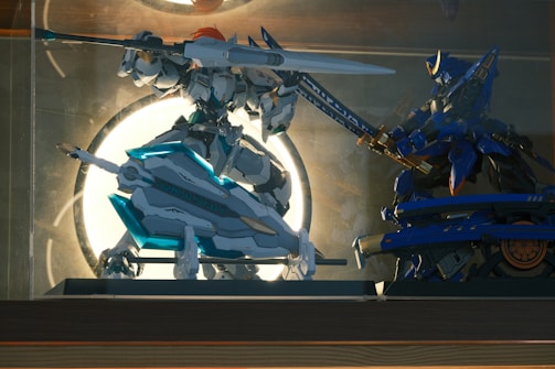 Two intricately designed robot figures engage in a dynamic pose within a glass display case. The left figure is predominantly white with turquoise accents, holding a large spear and shield, and features an orange element on its head. The right figure is primarily blue and gold, wielding a sword, poised in a combat stance. The background is illuminated by a bright circular light, creating a dramatic silhouette effect.