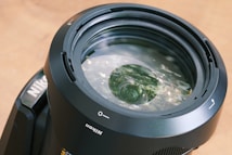 Close-up view of a camera lens with clear reflections and intricate glass elements. The lens appears robust, with labels and markings indicating orientation and brand. The surface is glossy, with subtle hints of ambient lighting reflected off the glass.