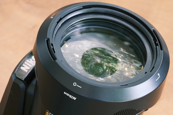 Close-up view of a camera lens with clear reflections and intricate glass elements. The lens appears robust, with labels and markings indicating orientation and brand. The surface is glossy, with subtle hints of ambient lighting reflected off the glass.