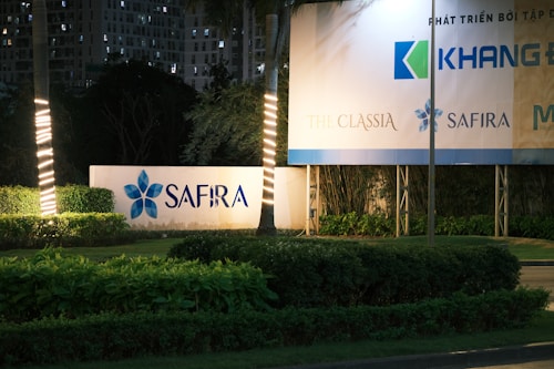 A well-lit sign displays the word 'SAFIRA' among neatly trimmed plants and landscaped greenery. Tall buildings are visible in the background, partially obscured by trees. Illuminated light wraps spiral around nearby palm trees, adding a decorative element to the scene.