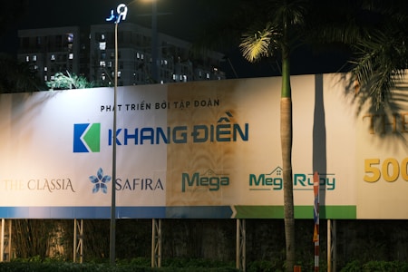 A nighttime scene featuring a large billboard displaying various real estate project logos and names. The billboard includes logos for 'Khang Dien,' 'The Classia,' 'Safira,' 'Mega Residences,' and 'Mega Ruby.' The setting is outdoors with a streetlight illuminating part of the billboard, and an apartment building is visible in the background. Palm trees and street signs are present in the foreground.