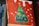 A canvas painting featuring a stylized green Christmas tree adorned with colorful decorations like fruits and flowers. The background is painted red with white spots, resembling snow. The artwork is propped up against a stack of books, with the spines partially visible.