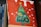 A canvas painting featuring a stylized green Christmas tree adorned with colorful decorations like fruits and flowers. The background is painted red with white spots, resembling snow. The artwork is propped up against a stack of books, with the spines partially visible.