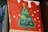 A canvas painting featuring a stylized green Christmas tree adorned with colorful decorations like fruits and flowers. The background is painted red with white spots, resembling snow. The artwork is propped up against a stack of books, with the spines partially visible.