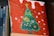 A canvas painting featuring a stylized green Christmas tree adorned with colorful decorations like fruits and flowers. The background is painted red with white spots, resembling snow. The artwork is propped up against a stack of books, with the spines partially visible.
