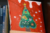 A canvas painting featuring a stylized green Christmas tree adorned with colorful decorations like fruits and flowers. The background is painted red with white spots, resembling snow. The artwork is propped up against a stack of books, with the spines partially visible.