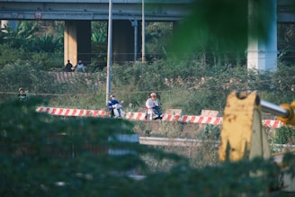 Several people are by a road secured with orange and white striped barriers. Two individuals are seated on motorbikes, wearing helmets. Dense vegetation surrounds the area, and an elevated highway is visible in the background.