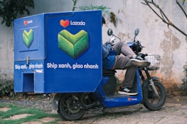 A delivery person is reclining on a blue scooter with a large Lazada-branded cargo box. The person is casually using a smartphone. The cargo box features a green and orange logo and the text 'Ship xanh, giao nhanh'. The setting appears to be an outdoor location with a worn plaster wall and some grass and stones on the ground.
