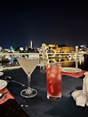 A nighttime outdoor dining setup overlooking a cityscape with lit buildings in the background. The foreground features a cocktail in a martini glass and a red drink in a tall glass with ice and a straw, placed on a dark tablecloth with cutlery and napkins nearby.