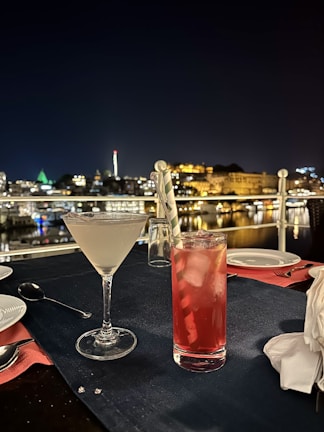 A nighttime outdoor dining setup overlooking a cityscape with lit buildings in the background. The foreground features a cocktail in a martini glass and a red drink in a tall glass with ice and a straw, placed on a dark tablecloth with cutlery and napkins nearby.