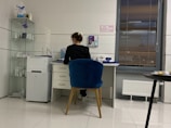 A person is seated at a desk in a modern, minimalist office space. They are working with various items such as papers and a cup on the desk. The room features white tiled floors and walls, with a blue cushioned chair. To the left, there is a shelving unit displaying several products, and a printer is positioned next to the desk. A large window with blinds is on the right side of the room.