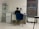 A person is seated at a desk in a modern, minimalist office space. They are working with various items such as papers and a cup on the desk. The room features white tiled floors and walls, with a blue cushioned chair. To the left, there is a shelving unit displaying several products, and a printer is positioned next to the desk. A large window with blinds is on the right side of the room.