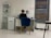 A person is seated at a desk in a modern, minimalist office space. They are working with various items such as papers and a cup on the desk. The room features white tiled floors and walls, with a blue cushioned chair. To the left, there is a shelving unit displaying several products, and a printer is positioned next to the desk. A large window with blinds is on the right side of the room.