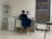 A person is seated at a desk in a modern, minimalist office space. They are working with various items such as papers and a cup on the desk. The room features white tiled floors and walls, with a blue cushioned chair. To the left, there is a shelving unit displaying several products, and a printer is positioned next to the desk. A large window with blinds is on the right side of the room.