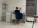 A person is seated at a desk in a modern, minimalist office space. They are working with various items such as papers and a cup on the desk. The room features white tiled floors and walls, with a blue cushioned chair. To the left, there is a shelving unit displaying several products, and a printer is positioned next to the desk. A large window with blinds is on the right side of the room.