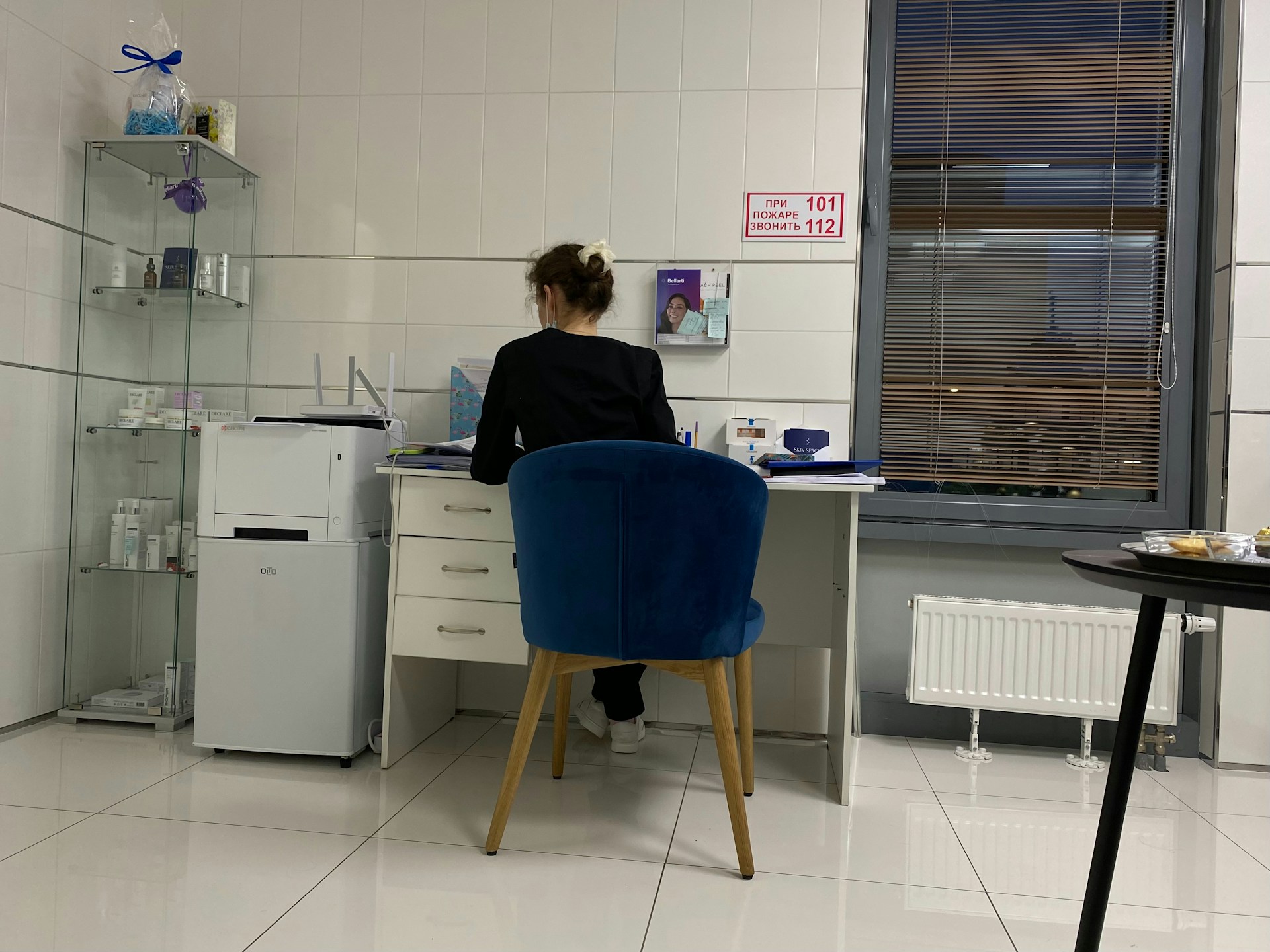 A person is seated at a desk in a modern, minimalist office space. They are working with various items such as papers and a cup on the desk. The room features white tiled floors and walls, with a blue cushioned chair. To the left, there is a shelving unit displaying several products, and a printer is positioned next to the desk. A large window with blinds is on the right side of the room.