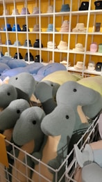 A collection of plush dinosaur toys with green and beige colors are placed in a white wire basket in front of a display of various bucket hats. The hats are neatly organized on white shelves against a yellow wall, featuring multiple colors including black, blue, and cream.