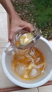 A hand is holding a small, clear pitcher containing a yellow, buttery substance being poured into a white bowl. The bowl contains yellow liquid and several large ice cubes. It is placed on a tiled surface with some grass visible in the background.