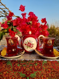 A decorative red teapot adorned with floral patterns is placed alongside two filled tea glasses on a tray. The background includes vibrant red bougainvillea flowers, and the setting appears to be outdoors, suggesting a warm and inviting environment.