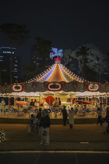 A brightly lit carousel with decorative lights and a Pegasus ornament on top, surrounded by people enjoying the ride at night. The background features trees and tall buildings with lights.