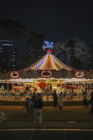 A brightly lit carousel with decorative lights and a Pegasus ornament on top, surrounded by people enjoying the ride at night. The background features trees and tall buildings with lights.