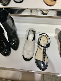 A display of fashionable shoes including a pair of black boots with shiny toes, a white sandal with a floral design on top, and a black sandal with branded straps. The shoes are arranged on a reflective white shelf with some metal elements.