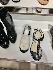A display of fashionable shoes including a pair of black boots with shiny toes, a white sandal with a floral design on top, and a black sandal with branded straps. The shoes are arranged on a reflective white shelf with some metal elements.