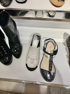 A display of fashionable shoes including a pair of black boots with shiny toes, a white sandal with a floral design on top, and a black sandal with branded straps. The shoes are arranged on a reflective white shelf with some metal elements.