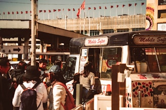 A bustling street scene featuring a mobile ice cream van with the words 'Mobile Softee' on it, surrounded by a group of people standing and waiting. The background shows a building with numerous small red flags lining the roof. The setting appears lively and busy, typical of an urban environment.