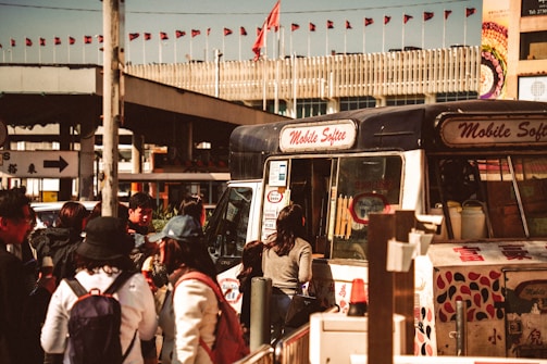 A bustling street scene featuring a mobile ice cream van with the words 'Mobile Softee' on it, surrounded by a group of people standing and waiting. The background shows a building with numerous small red flags lining the roof. The setting appears lively and busy, typical of an urban environment.