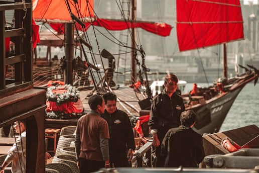 Several people are standing on a traditional wooden ship with vibrant red sails. The ship is docked by the water, and a few decorations and ropes are visible on the deck. The atmosphere appears busy and lively, with some individuals appearing to be engaged in conversation.