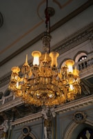 An ornate crystal chandelier hangs from an intricately decorated ceiling inside a lavish interior space. The chandelier is illuminated, casting a warm glow that highlights its detailed design. The background features decorative moldings, arched openings, and sculpted figures, contributing to an opulent and grand atmosphere.