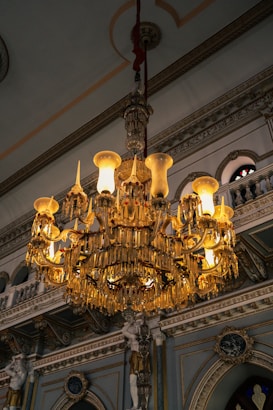 An ornate crystal chandelier hangs from an intricately decorated ceiling inside a lavish interior space. The chandelier is illuminated, casting a warm glow that highlights its detailed design. The background features decorative moldings, arched openings, and sculpted figures, contributing to an opulent and grand atmosphere.