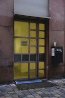 A modern door with a grid design, featuring rectangular yellow glass panels set in a metal frame. It is flanked by dark stone walls and sits above a cobblestone path. A black mailbox with the number 23 is mounted on the right side next to a takeaway coffee cup.