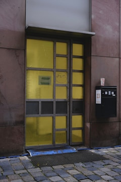 A modern door with a grid design, featuring rectangular yellow glass panels set in a metal frame. It is flanked by dark stone walls and sits above a cobblestone path. A black mailbox with the number 23 is mounted on the right side next to a takeaway coffee cup.