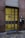 A modern door with a grid design, featuring rectangular yellow glass panels set in a metal frame. It is flanked by dark stone walls and sits above a cobblestone path. A black mailbox with the number 23 is mounted on the right side next to a takeaway coffee cup.