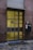 A modern door with a grid design, featuring rectangular yellow glass panels set in a metal frame. It is flanked by dark stone walls and sits above a cobblestone path. A black mailbox with the number 23 is mounted on the right side next to a takeaway coffee cup.
